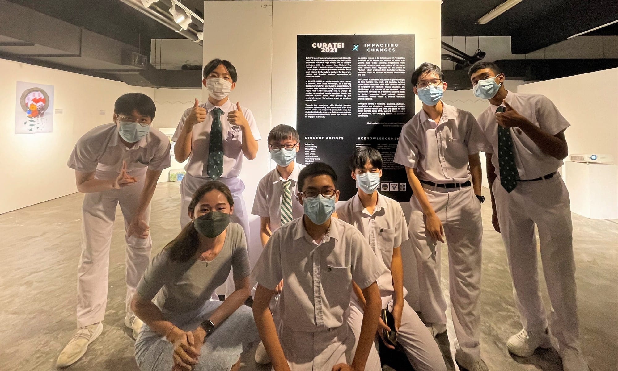 Group of masked students posing in front of a Curate 2021 Impacting Changes exhibition board in a gallery.